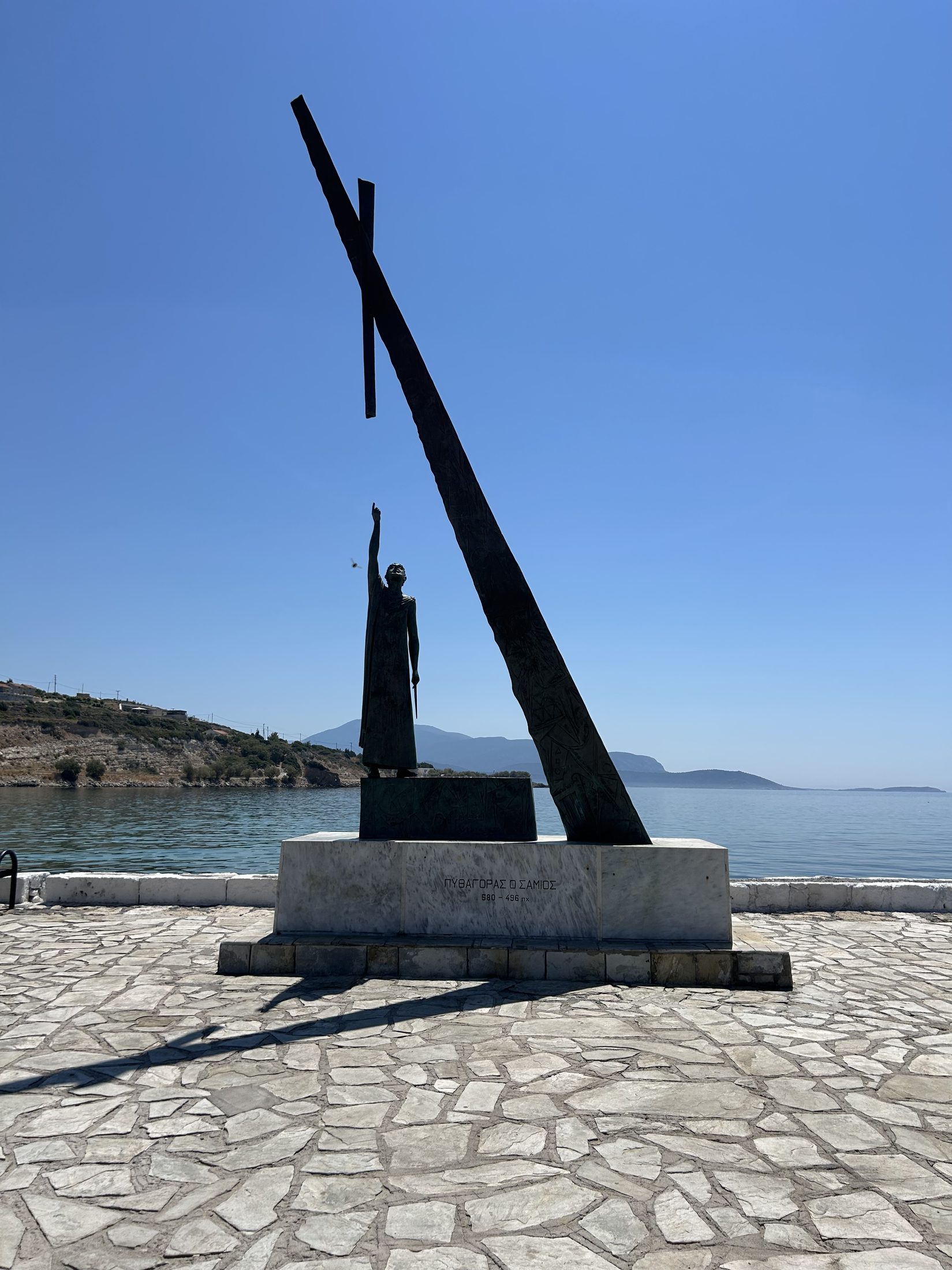 Statue of Pythagoras of Samos on the harbourfront, arm raised toward the sea, beside a large bronze right-triangle sculpture.
