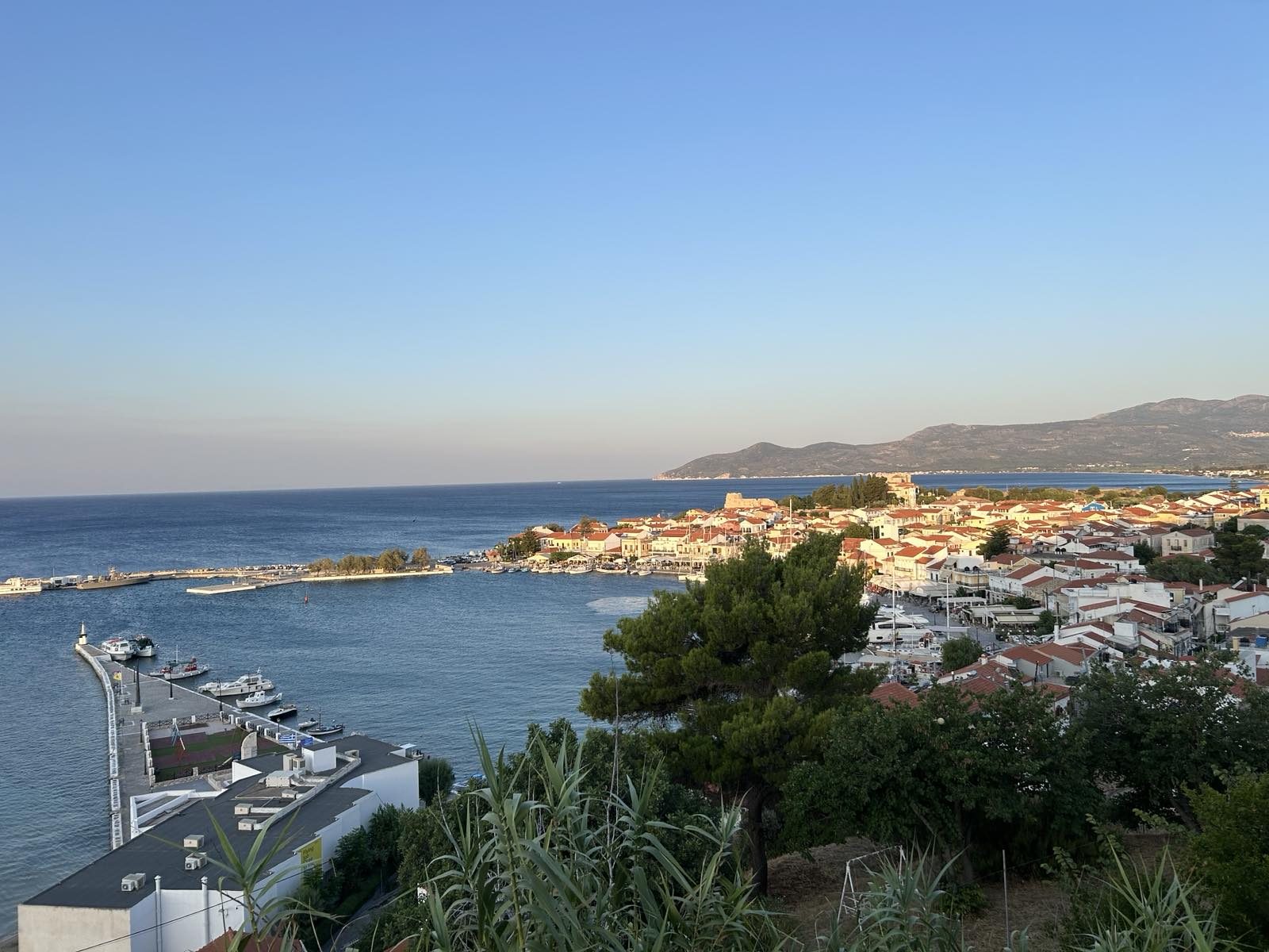 Aerial view of Samos Town and its harbour, whitewashed houses with terracotta roofs along the Aegean coastline, mountains in the distance.